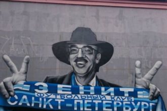 Известный артист изображен в черной шляпе, очках и шарфом с надписью «Футбольный клуб «Зенит» Санкт-Петербург» в руках.