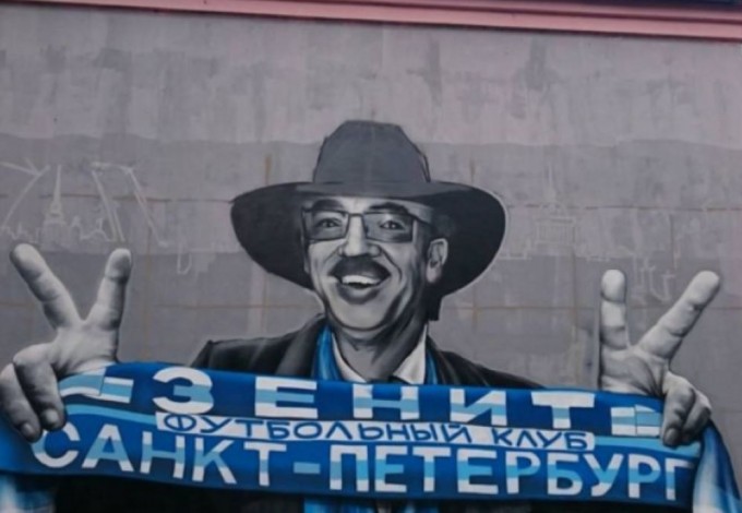 Известный артист изображен в черной шляпе, очках и шарфом с надписью «Футбольный клуб «Зенит» Санкт-Петербург» в руках.